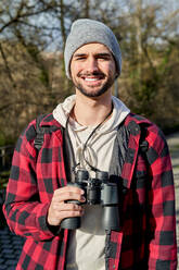 Lächelnder bärtiger männlicher Reisender mit Fernglas in der Hand, der interessiert in die Kamera schaut, während er auf einem Weg in der Nähe eines Zauns in der Stadt steht - ADSF39937