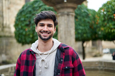 Positive bearded male traveler in casual clothes looking at camera with smile while standing near ancient building and fountain during trip in old town - ADSF39941