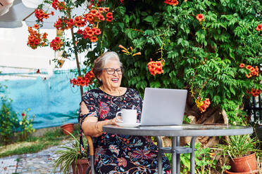 Positive ältere Dame in legerem Outfit und Brille sitzt im Stuhl am Tisch beim Surfen auf Netbook in der Nähe von Tasse auf Untertasse und Grünpflanzen mit Blumen im Hof im Tageslicht - ADSF39944