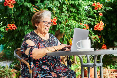 Positive ältere Dame in legerem Outfit und Brille sitzt im Stuhl am Tisch beim Surfen auf Netbook in der Nähe von Tasse auf Untertasse und Grünpflanzen mit Blumen im Hof im Tageslicht - ADSF39945