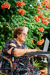 Konzentrierte ältere Dame in legerem Outfit und Brille sitzt auf einem Stuhl am Tisch und surft auf einem Netbook in der Nähe einer Tasse auf einer Untertasse und Grünpflanzen mit Blumen im Garten bei Tageslicht - ADSF39947