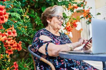 Ältere Frau mit Brille und Freizeitkleidung, die ein Mobiltelefon benutzt, während sie auf einem Stuhl in der Nähe eines Tisches mit Laptop im Garten in der Nähe von Blumen und Pflanzen im Sommer sitzt - ADSF39950