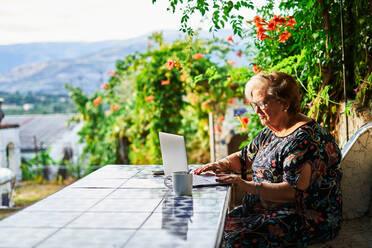 Positive ältere Dame in legerem Outfit und Brille sitzt im Stuhl am Tisch beim Surfen auf Netbook in der Nähe von Tasse auf Untertasse und Grünpflanzen mit Blumen im Hof im Tageslicht - ADSF39954