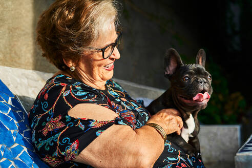 Smiling aged lady in eyeglasses sitting with French Bulldog on bench in daylight in yard - ADSF39955