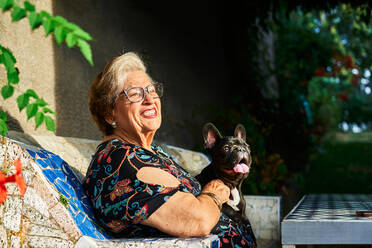 Smiling aged lady in eyeglasses sitting with French Bulldog on bench in daylight in yard - ADSF39956