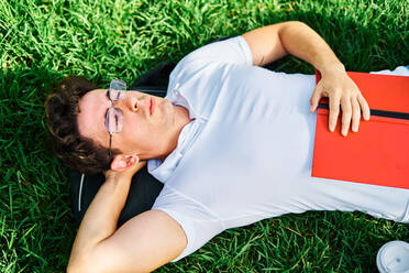 From above of male student lying on grass with closed eyes and having rest in campus on sunny day - ADSF39960