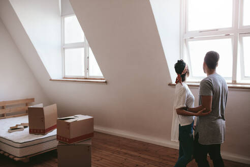 Young man and woman standing by window in new home. Young couple moving in new house with cardboard boxes. - JLPSF27061