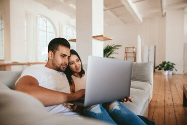 Liebevolles junges Paar sitzt auf dem Sofa und benutzt einen Laptop. Junger Mann und Frau entspannen sich auf der Couch mit einem Laptop zu Hause. - JLPSF27064