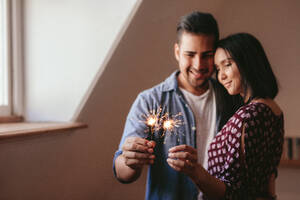 Glücklicher junger Mann und Frau halten Wunderkerzen zu Hause. Schönes junges Paar, das mit Wunderkerzen in einem Haus feiert. - JLPSF27069
