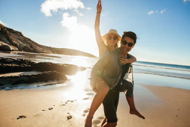 Porträt eines gutaussehenden jungen Mannes, der seine hübsche Freundin auf dem Rücken trägt, während ein Paar den Strandurlaub genießt. - JLPSF27074