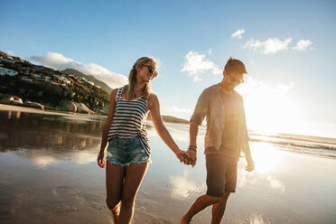 Außenaufnahme eines jungen Paares, das am Meer spazieren geht und sich an den Händen hält. Junger Mann und Frau, die zusammen am Strand spazieren gehen. - JLPSF27077