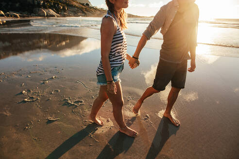Außenaufnahme eines jungen Paares, das am Meer spazieren geht und sich an den Händen hält. Junger Mann und Frau, die zusammen am Strand spazieren gehen. - JLPSF27081