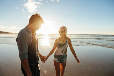 Außenaufnahme eines romantischen jungen Paares, das sich an den Händen hält und am Strand spazieren geht. Junger Mann und Frau gehen an einem Sommertag am Meer spazieren. - JLPSF27082