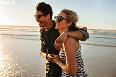 Außenaufnahme eines lächelnden jungen Paares, das am Strand spazieren geht. Junger Mann und Frau, die an einem Sommertag gemeinsam am Meer spazieren gehen. - JLPSF27085