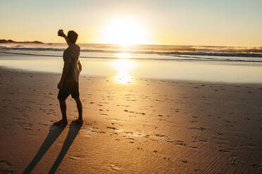 Rückansicht eines jungen Mannes, der ein Selfie bei Sonnenuntergang am Strand macht. Ein Mann, der ein Selbstporträt am Meeresufer macht. - JLPSF27093