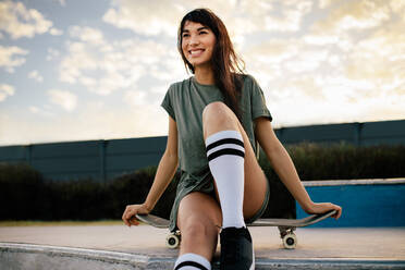 Attraktive Frau sitzt auf dem Skateboard und schaut lächelnd weg, während sie Zeit im Skatepark verbringt. Urbanes Mädchen entspannt sich im Skatepark. - JLPSF27098