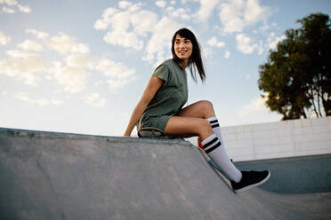 Weibliche Skateboarderin auf einer Rampe im Skatepark sitzend. Frau macht eine Pause nach dem Skateboardfahren im Park. - JLPSF27104