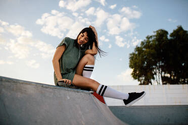Weibliche Skateboarderin, die sich im Skatepark amüsiert. Glückliche Frau sitzt auf einer Skateboard-Rampe im Freien und lächelt. - JLPSF27106