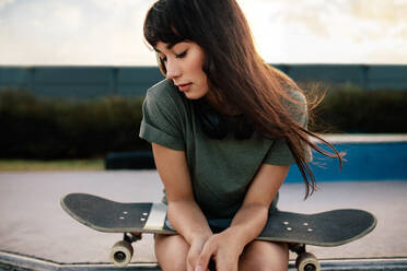 Junge Frau sitzt im Skatepark mit Skateboard und schaut nach unten. Junge Frau auf dem Skateboard entspannt sich im Skatepark. - JLPSF27114