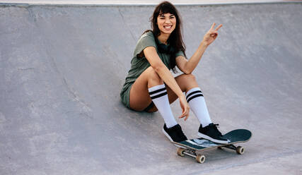 Porträt einer schönen jungen Frau, die auf einer Skateparkrampe mit einem Skateboard sitzt, lächelt und ein Friedenshandzeichen macht. - JLPSF27116