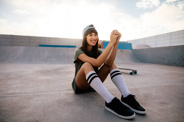 Fröhliche Skateboarderin, die im Skatepark sitzt, wegschaut und lächelt. Coole Frau, die sich im Freien im Skatepark entspannt. - JLPSF27120