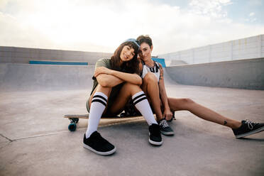 Two female skateboarders sitting on a long board at skate park. Woman friends hanging out together outdoors at skate park. - JLPSF27123