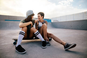 Two female friends enjoying spending time together at skate park. Woman skateboarders sitting on long board and looking at each other. - JLPSF27124