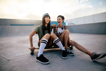 Städtische Mädchen im Skatepark mit Skateboard lachen und haben Spaß. Zwei Freundinnen sitzen auf einem langen Brett und lachen. - JLPSF27126