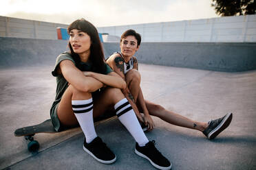 Zwei junge Frauen sitzen auf einem Longboard und schauen weg. Freundinnen sitzen entspannt im Skatepark. - JLPSF27128
