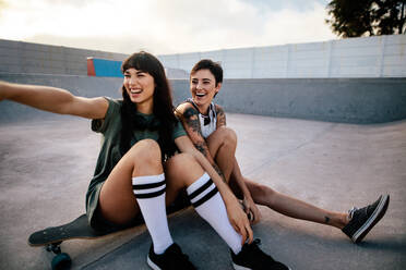 Zwei Skateboarderinnen sitzen auf einem langen Brett, wobei eine auf etwas Interessantes im Skatepark zeigt. - JLPSF27129