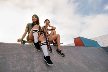 Zwei junge, urbane Mädchen im Skatepark, die lachen und Spaß haben. Zwei Freundinnen sitzen auf einem Longboard. - JLPSF27140