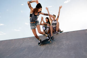 Skaterin fährt auf Skateboard mit Freundinnen, die auf der Rampe sitzen und jubeln. Weibliches Skateboarding im Skatepark mit jubelnden Freunden. - JLPSF27141