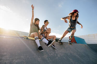 Zwei junge Frauen sitzen auf einer Skateboard-Rampe mit einem Mädchen, das Skateboarden übt. Skateboardfahrende Frau fährt Skateboard auf einer Skatepark-Rampe mit Freunden, die daneben sitzen. - JLPSF27143