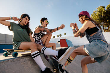 Drei junge Frauen, die sich im Skatepark amüsieren. Junge Frau beim Skateboarden mit Freunden auf der Rampe sitzend. - JLPSF27144