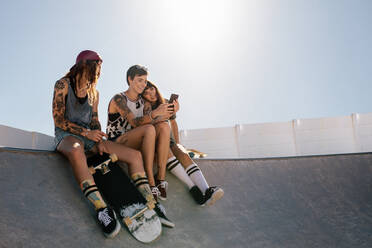 Group of female friends sitting on ramp at skate park and looking at mobile phone. Three women skaters using smart phone together at skate park. - JLPSF27162