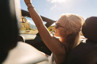 Schöne Frau genießt auf einer Autoreise. Frau sitzt auf dem Beifahrersitz eines Cabriolets und hält ihre Hand in die Luft. - JLPSF27189