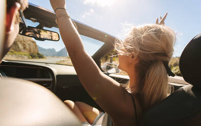 Frau genießt auf einer Autoreise mit Mann fahren Auto. Frau sitzt auf dem Vordersitz eines Cabriolets mit ihren Händen in der Luft erhoben, während ihr Freund Auto fahren. - JLPSF27190