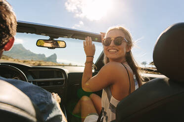 Pretty woman sitting in front passenger seat of a convertible car and looking back at camera. Smiling woman with her man traveling by a car on road trip. - JLPSF27191