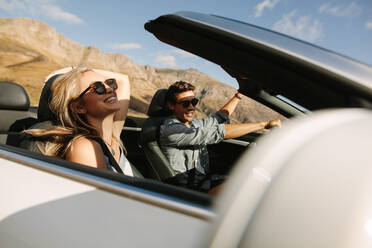Cheerful couple having fun on a road trip. Young man driving convertible car with woman smiling on passenger seat. - JLPSF27195