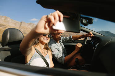 Paar auf Autoreise im Cabrio, das Selfie macht. Frau spricht Selfie auf langer Fahrt mit ihrem Mann, der das Auto fährt. - JLPSF27197