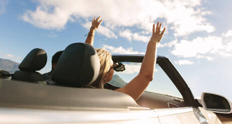 Rear view shot of a man and woman driving in a convertible car with raised hands. Couple enjoying themselves on a road trip. - JLPSF27199