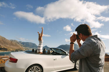 Man taking pictures of a woman standing in a convertible car. Couple taking pictures on road trip. - JLPSF27203