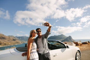 Ein Mann macht ein Selfie mit seiner Freundin auf einer Autoreise. Ein Paar steht bei seinem Auto und macht ein Selfie mit einem Mobiltelefon im Freien. - JLPSF27208
