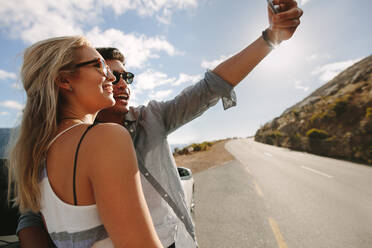 Ein Paar macht ein Selfie mit einem Smartphone und lächelt, während es in der Nähe des Autos steht. Ein Mann und eine Frau posieren für ein Selbstporträt neben ihrem Auto auf einer Autoreise. - JLPSF27210