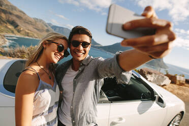 Ein Mann macht ein Selfie mit seiner Frau auf einer Autoreise. Ein Paar steht neben seinem Cabrio und macht ein Selfie mit einem Handy. - JLPSF27213