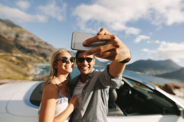 Schönes Paar steht zusammen und lehnt sich an ihr Auto, während sie ein Selfie machen. Fokus auf die Hand eines Mannes, der sein Handy hält, um ein Selfie mit seiner Freundin zu machen. - JLPSF27214