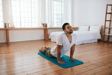 Innenaufnahme eines gutaussehenden jungen Mannes, der Yoga praktiziert. Fitness-Mann meditiert mit geschlossenen Augen, während er die Kobra-Pose im Wohnzimmer einnimmt. - JLPSF27229