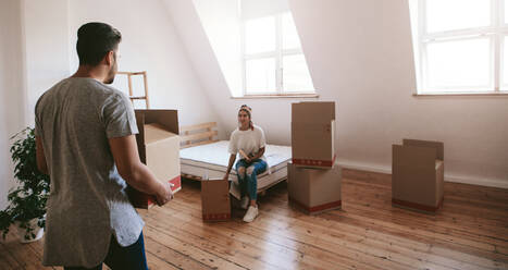 Indoor shot of young woman sitting on bed with man carrying box in new house. Young couple moving into new home. - JLPSF27233