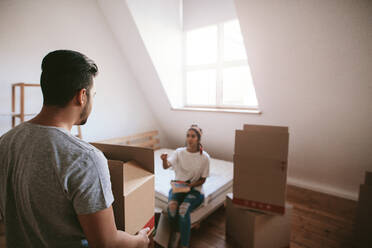 Rückansicht eines jungen Mannes, der eine Kiste trägt, mit einer Frau, die auf einem Bett im Hintergrund sitzt. Ein junges Paar zieht in ein neues Haus ein. - JLPSF27234