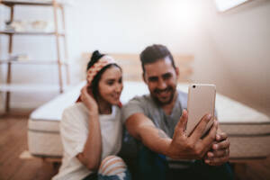 Aufnahme eines jungen Paares, das zu Hause sitzt und ein Selfie mit dem Mobiltelefon macht, wobei das Smartphone in den Händen des Mannes im Mittelpunkt steht. - JLPSF27237